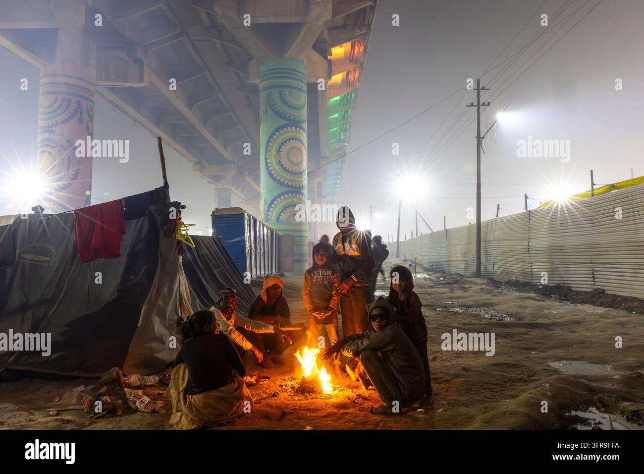Famiglia e bambini si riscaldano le mani attorno a un piccolo fuoco accanto a un sovrappasso con pilastri dipinti durante il Maha Kumbh Mela, Uttar Pradesh, Prayagraj, India Foto Stock