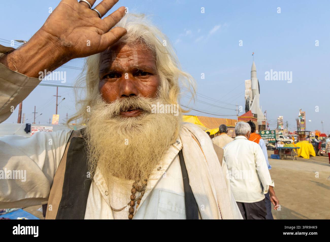 Uomo anziano barbuto solleva la mano alla fronte con un modello di razzo e persone dietro di lui alla Maha Kumbh Mela, Uttar Pradesh, Prayagraj, India Foto Stock