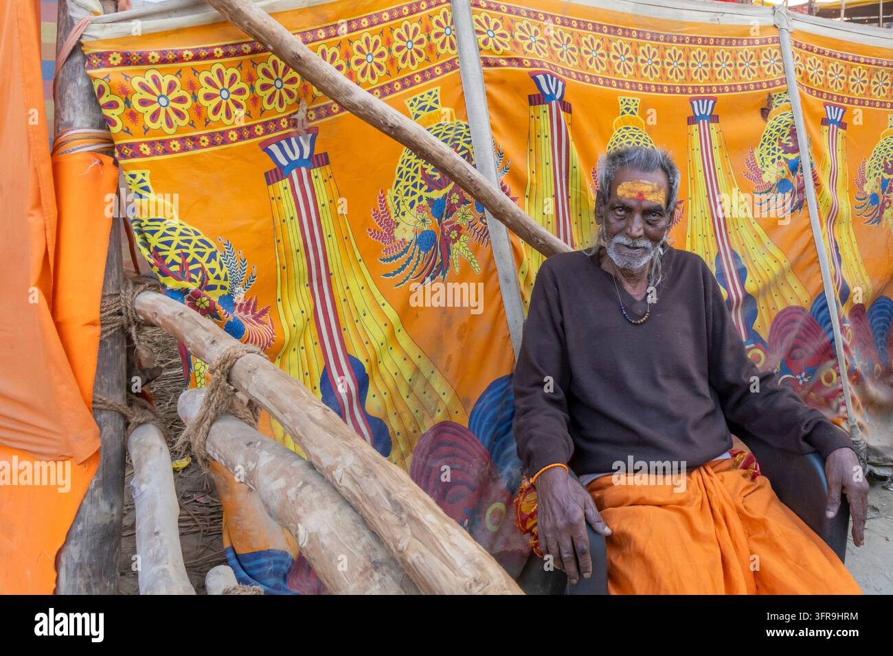 Uomo anziano con un tilak giallo sulla fronte seduto in abiti arancioni davanti a una tenda colorata al Maha Kumbh Mela, Uttar Pradesh, Prayagraj, India Foto Stock