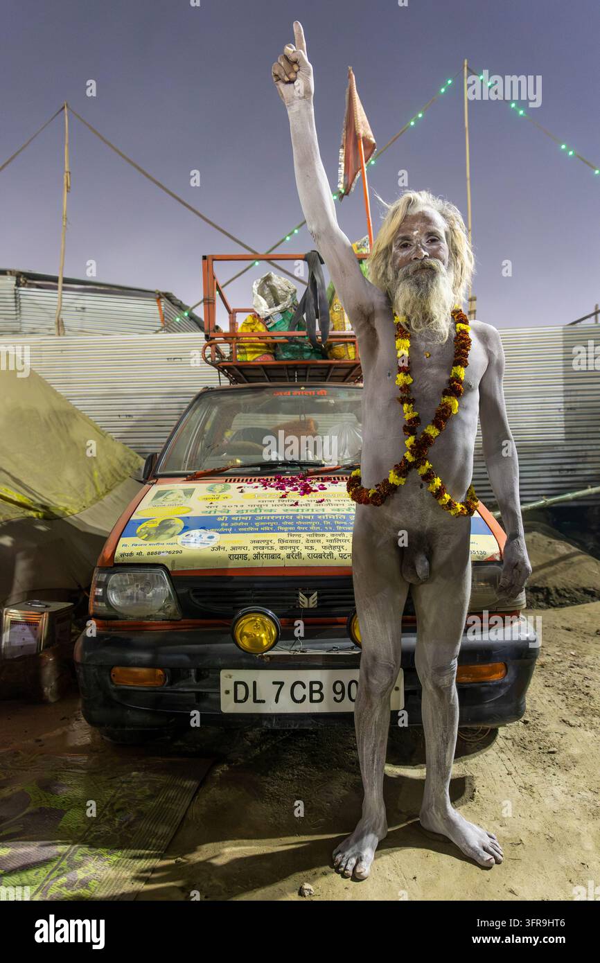 Naga Sadhu coperto di cenere indica il cielo indossando una ghirlanda di fiori di garofano vicino a un'auto al Maha Kumbh Mela, Uttar Pradesh, Prayagraj, India Foto Stock