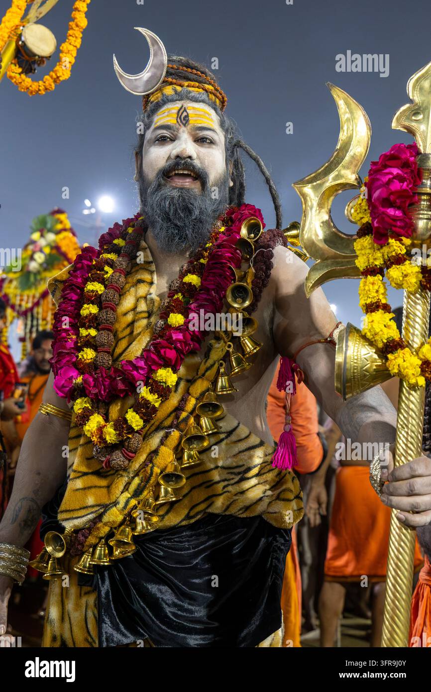 Naga Sadhu sorridente con copricapo a mezzaluna che tiene un tridente dorato con ghirlande di fiori alla Maha Kumbh Mela, Uttar Pradesh, Prayagraj, India Foto Stock