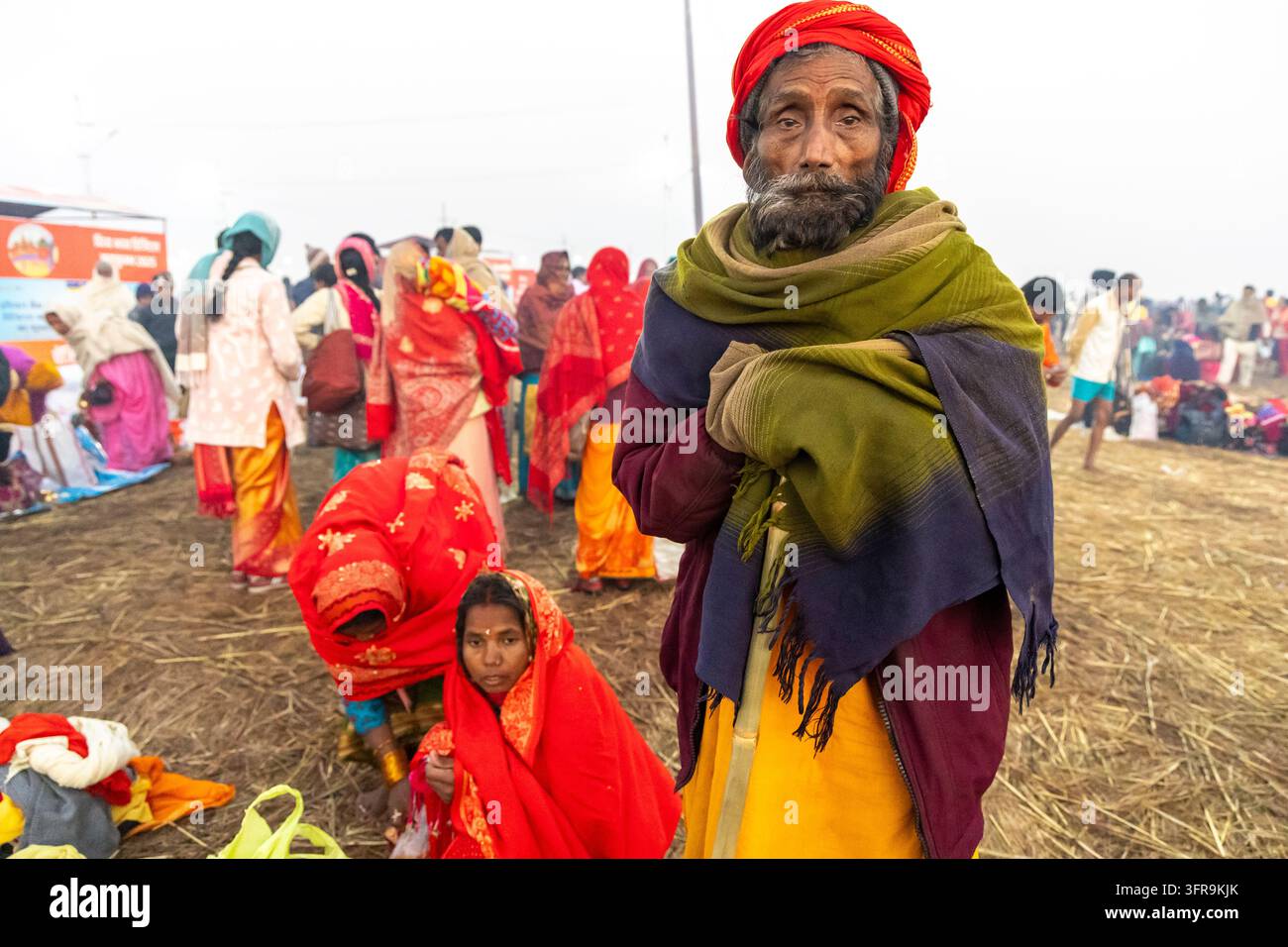 Uomo anziano con la barba, turban rosso e scialle verde, si trova tra donne sedute in sari rossi alla Maha Kumbh Mela, Uttar Pradesh, Prayagraj, India Foto Stock