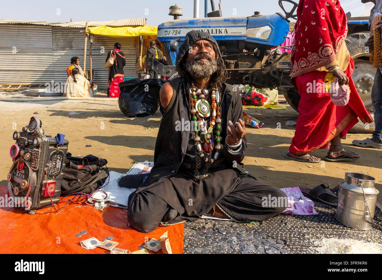 Uomo barbuto in nero con perline seduto per terra che mendica accanto a un trattore al Maha Kumbh Mela, Uttar Pradesh, Prayagraj, India Foto Stock