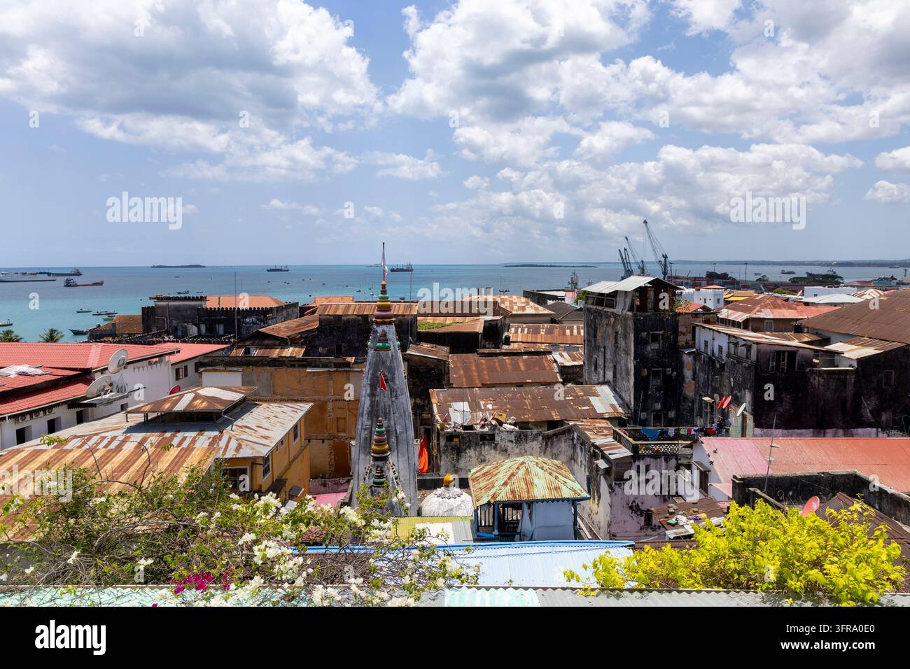 Vista su tetti di lamiera arrugginiti e vecchi edifici con una guglia di tempio indù e il porto con navi e gru in lontananza sotto un cielo nuvoloso e soleggiato, Zanzibar Foto Stock