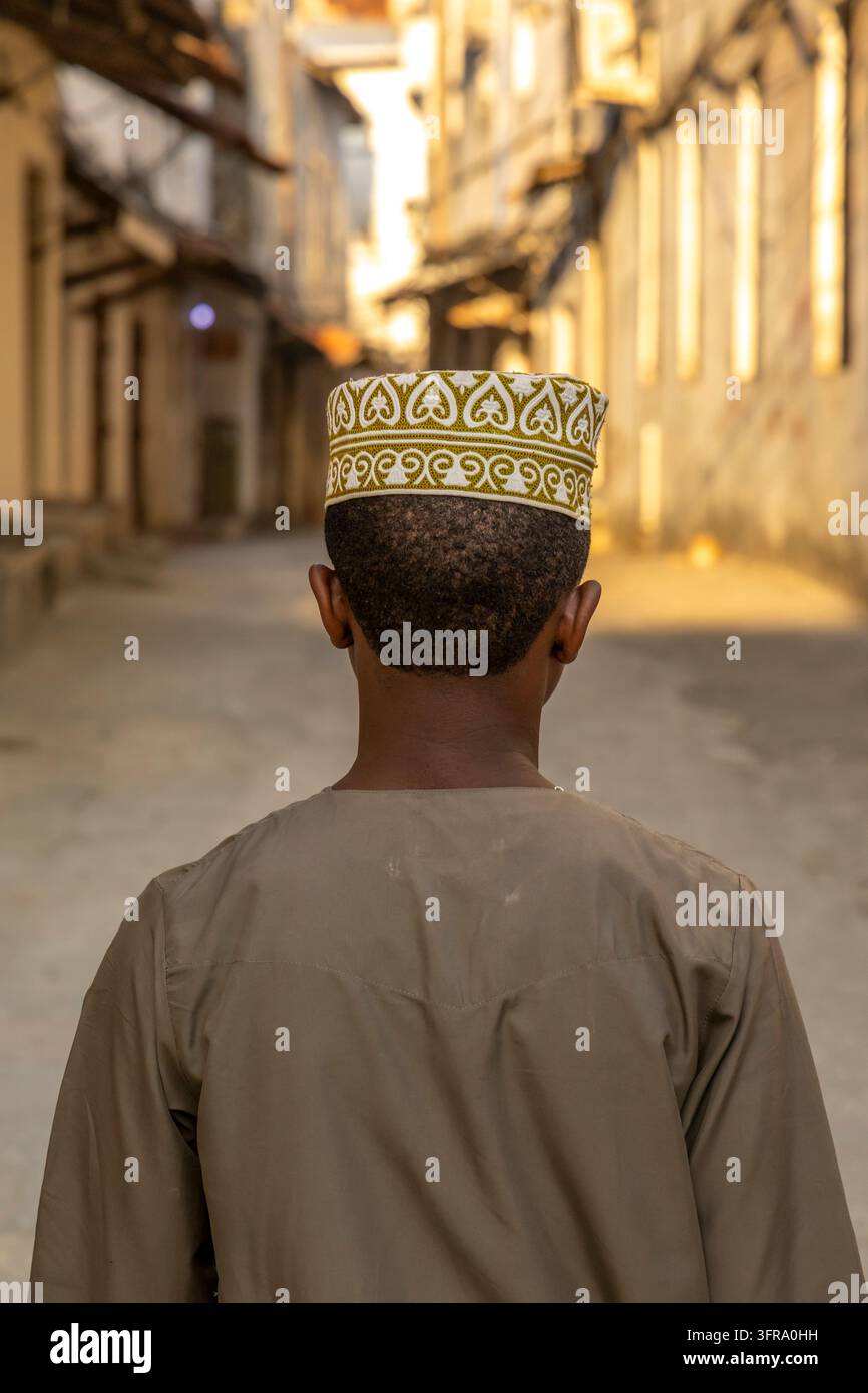 Vista posteriore di un ragazzo che indossa un cappello kofia ricamato verde e bianco e una veste marrone mentre cammina lungo una stretta via soleggiata tra vecchi edifici, Zanzibar Foto Stock