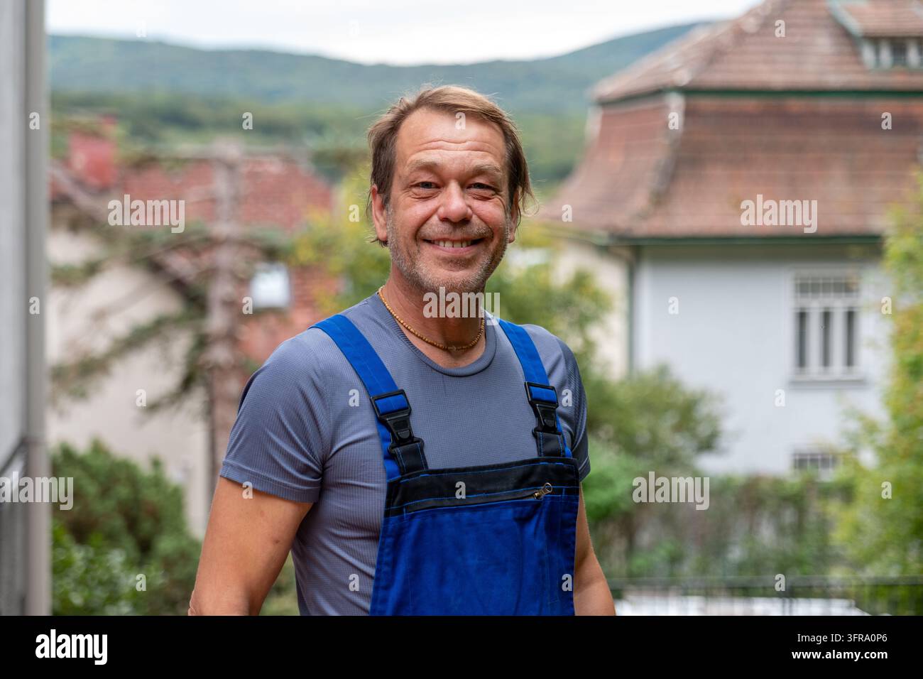 Un uomo di mezza età sorridente si trova all'aperto davanti a case di villaggio e colline sfocate. Foto Stock