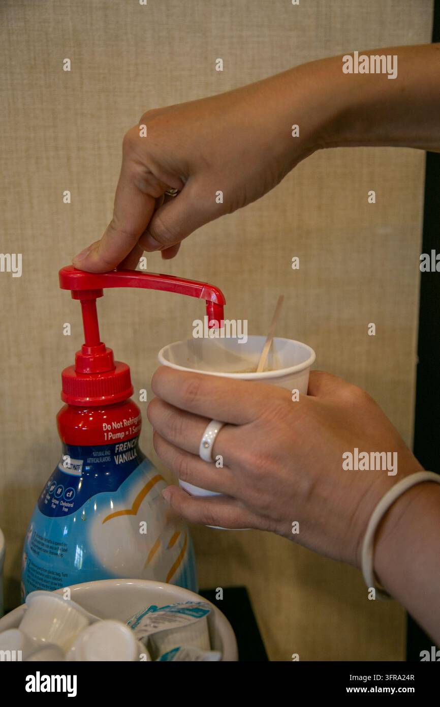Primo piano delle mani di una persona che eroga latte di caffè liquido da una grande bottiglia a pompa commerciale in una piccola tazza di carta bianca. Bevande self-service Foto Stock