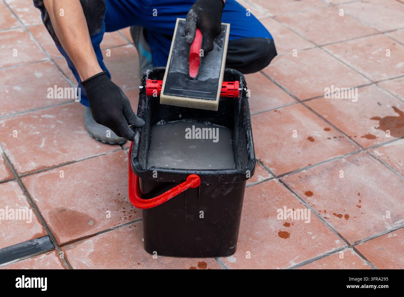 Un operaio guantato pulisce un pavimento in piastrelle di terracotta bagnato con una spugna e un secchio. Foto Stock