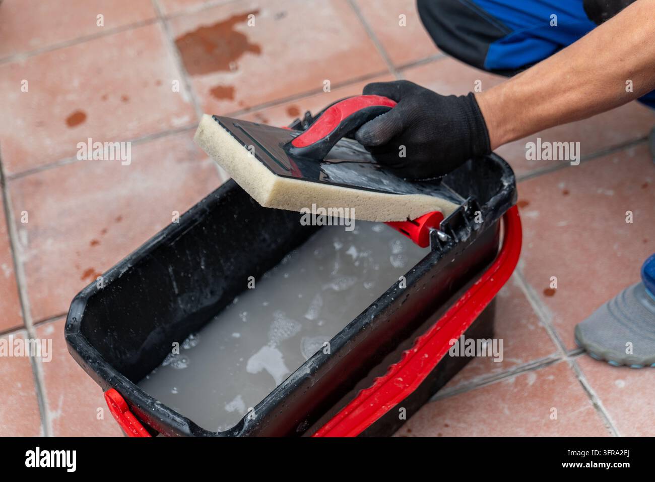Una mano guantata risciacqua una spugna per mocio in un secchio su un pavimento piastrellato. Foto Stock