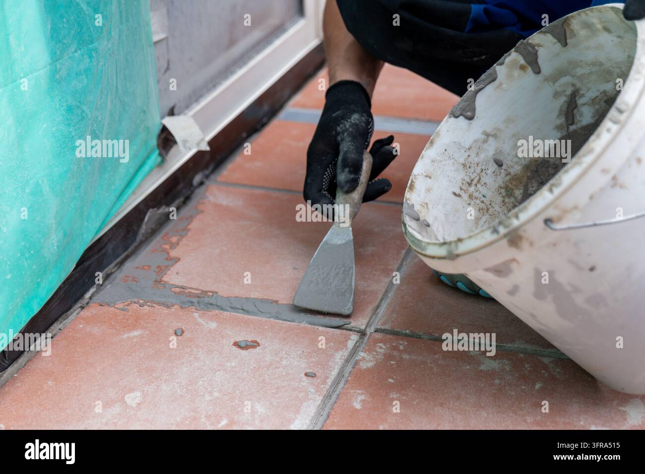 Un operaio guantato applica una malta grigia tra le piastrelle di terracotta con una spatola. Foto Stock