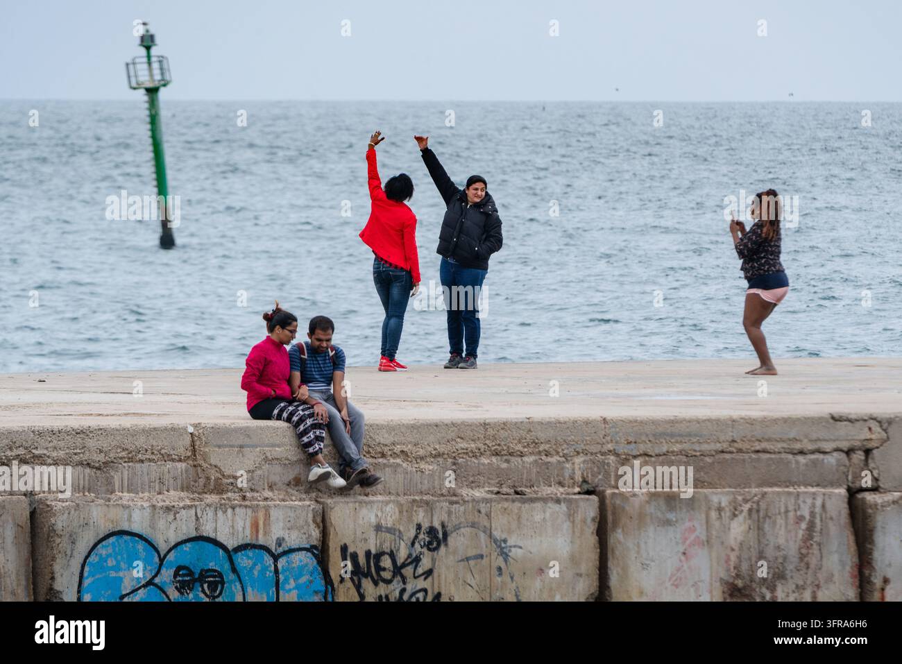 AMICI CHE SI DIVERTISCONO E FANNO FOTO SU UN MOLO, PLAYA DE LA BARCELONETA REPORTAGE, BARCELLONA, SPAGNA: Un gruppo di amici si diverte su un molo di cemento in riva al mare, con una donna che scatta una foto ai suoi due amici che posano mentre una coppia è seduta nelle vicinanze. INFO: La spiaggia di Barceloneta è la destinazione balneare più famosa e vivace di Barcellona. Famosa per le sue sabbie inclinate e il vivace viale alberato, unisce prendere il sole e sport acquatici con bar e ristoranti animati sul lungomare, moli di cemento e zone fitness. Foto Stock