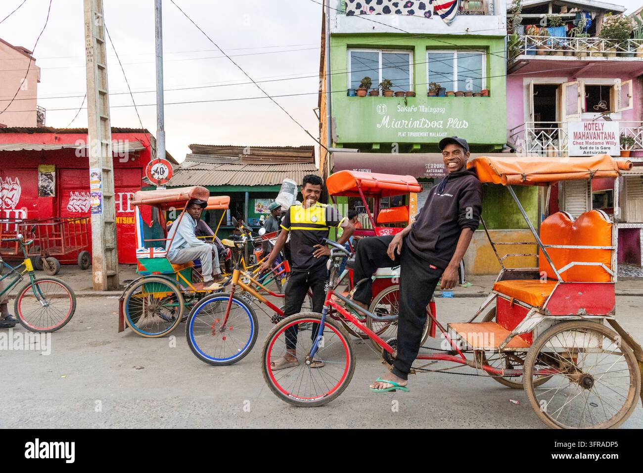 Tiratori di risciò che posano e sorridono accanto ai loro taxi biciclette su una strada colorata fiancheggiata da negozi e hotel, Vakinankaratra, Antsirabe, Madagascar Foto Stock