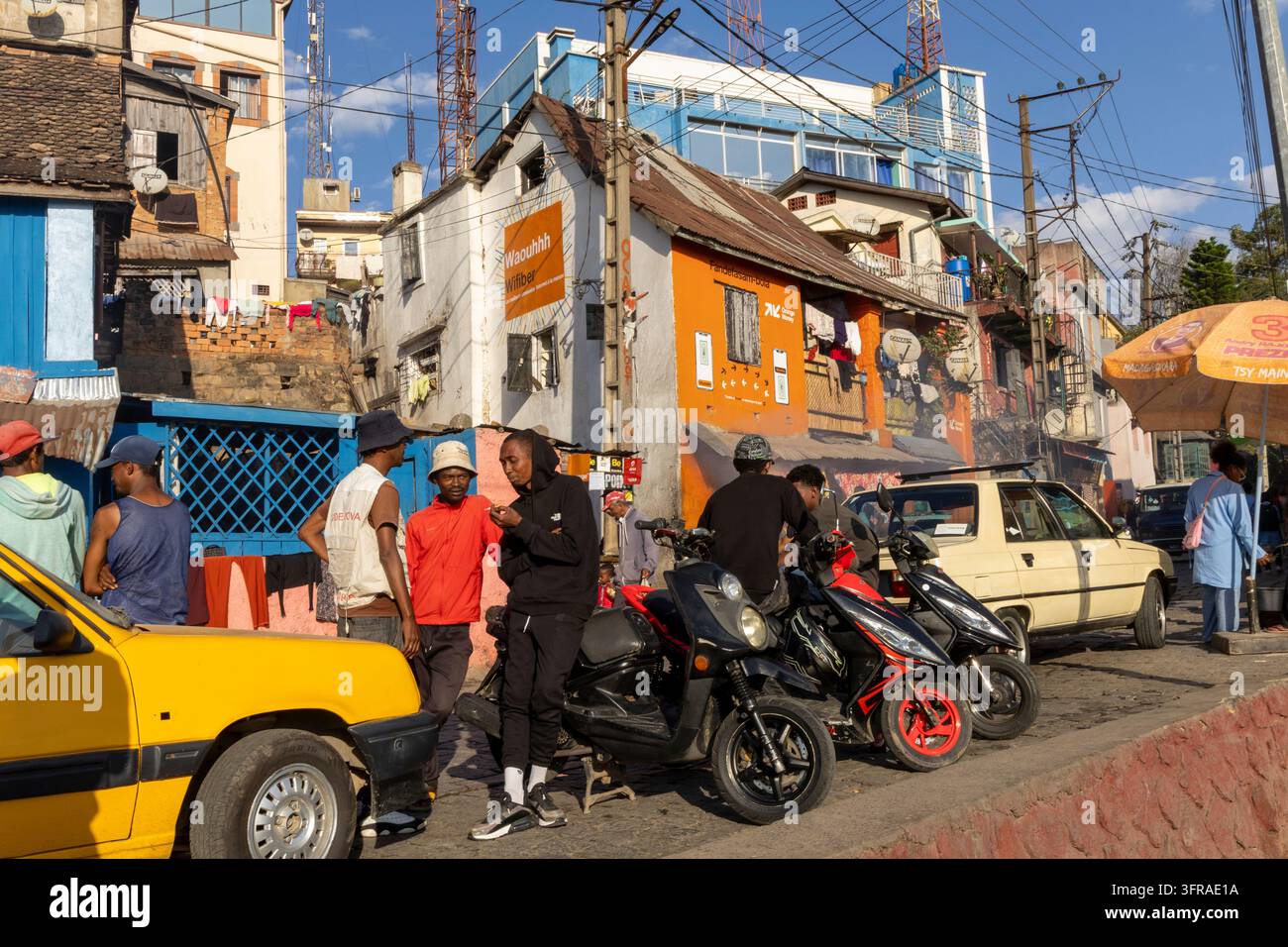 Donna che passa davanti a facciate di negozi dipinte con colori vivaci lungo una strada acciottolata fiancheggiata da pubblicità per il denaro mobile, Analamanga, Antananarivo, Madagascar Foto Stock