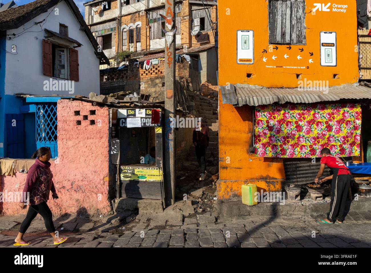 Donna che passa davanti a un angolo di strada colorato dove un uomo griglia cibo accanto a facciate di negozi dipinte, Analamanga, Antananarivo, Madagascar Foto Stock