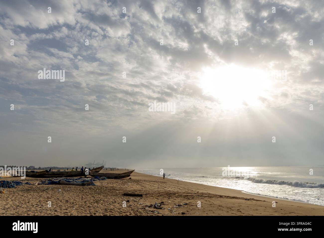 Piroge di legno appoggiate su una spiaggia sotto un cielo nuvoloso drammatico con gru portuali visibili in lontananza, Regione Marittima, Lomé, Togo Foto Stock