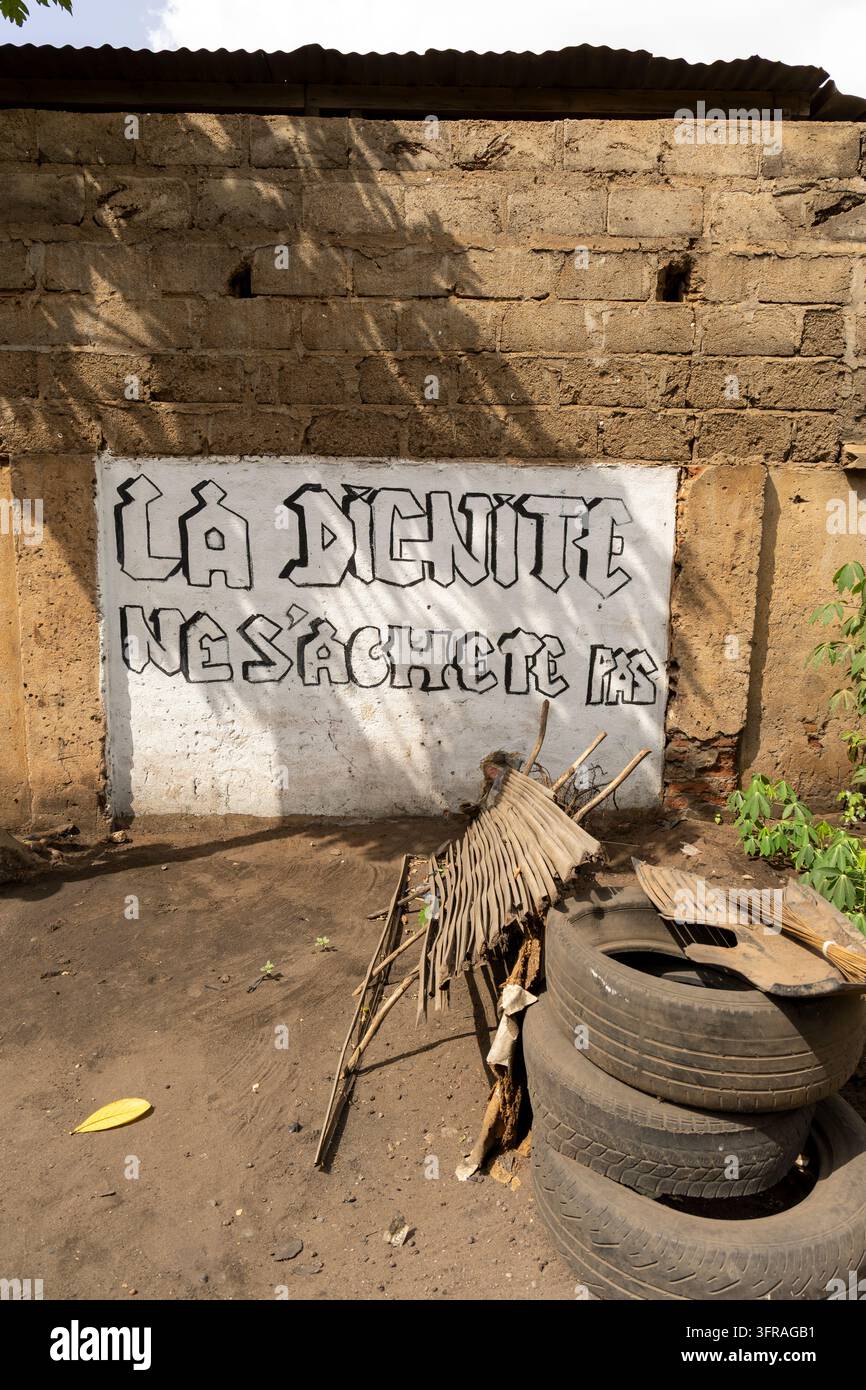 Un muro di graffiti con la scritta "la dignità non è in vendita" si erge sopra pneumatici vecchi impilati e costole di palma essiccate in un cortile sabbioso, Regione Marittima, Lomé, Togo Foto Stock