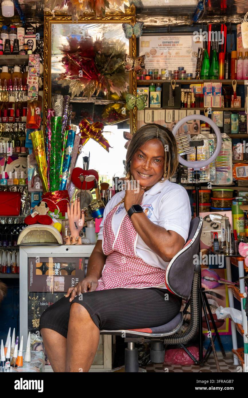 Una estetista posa per un ritratto all'interno del suo piccolo chiosco di salone per unghie e cosmetici decorato con uno specchio e un certificato, Regione Marittima, Lomé, Togo Foto Stock