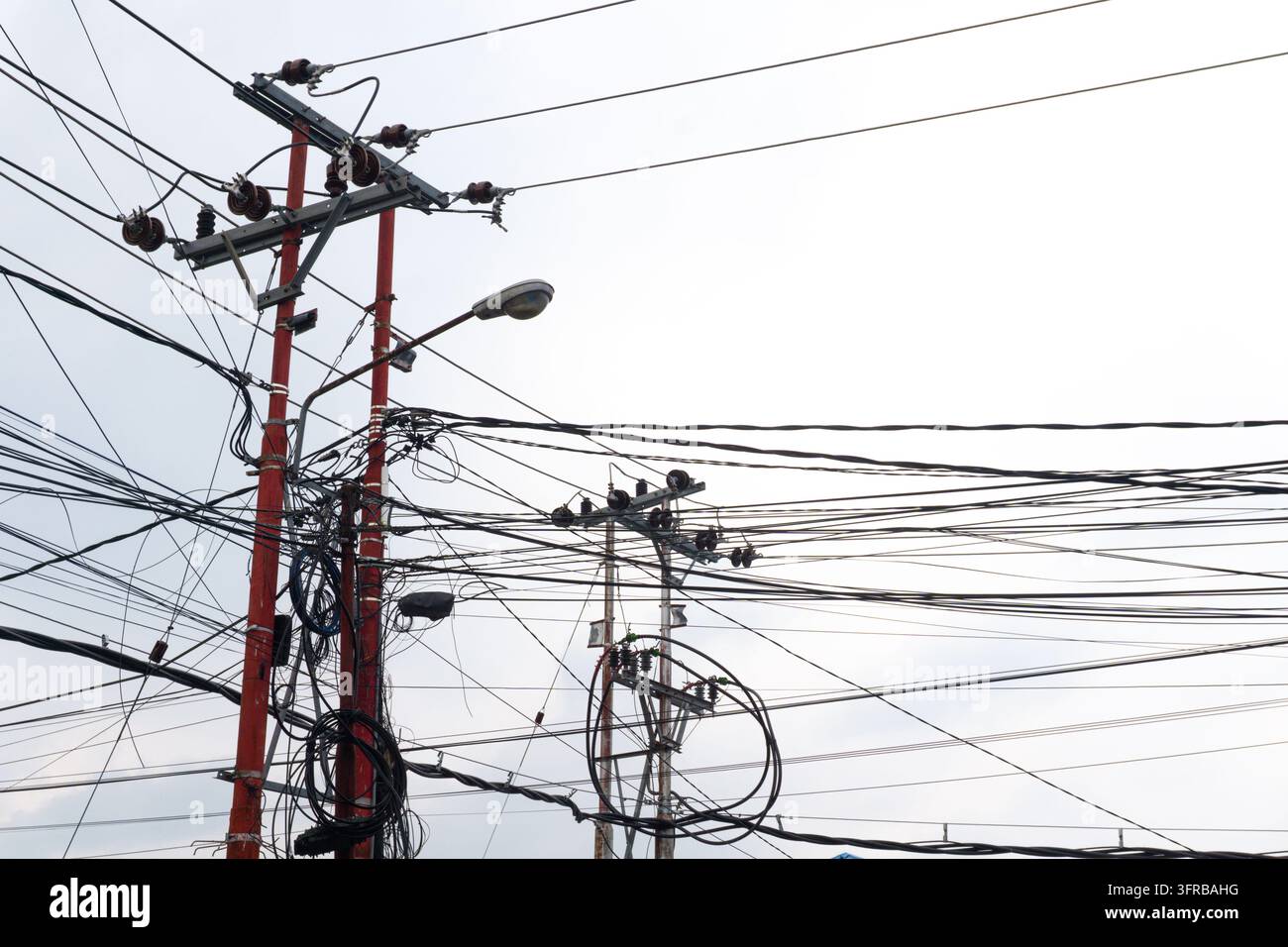 Cavi e fili elettrici ingarbugliati su pali dell'energia con isolatori e un lampione contro un cielo nuvoloso Foto Stock