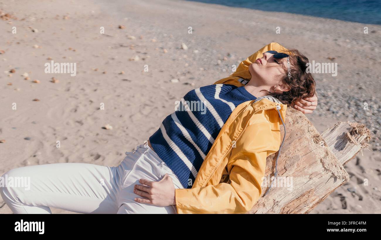 Donna in spiaggia che si rilassa, donna con impermeabile giallo e maglione a righe si appoggia su un pezzo di legno alla spiaggia soleggiata Foto Stock
