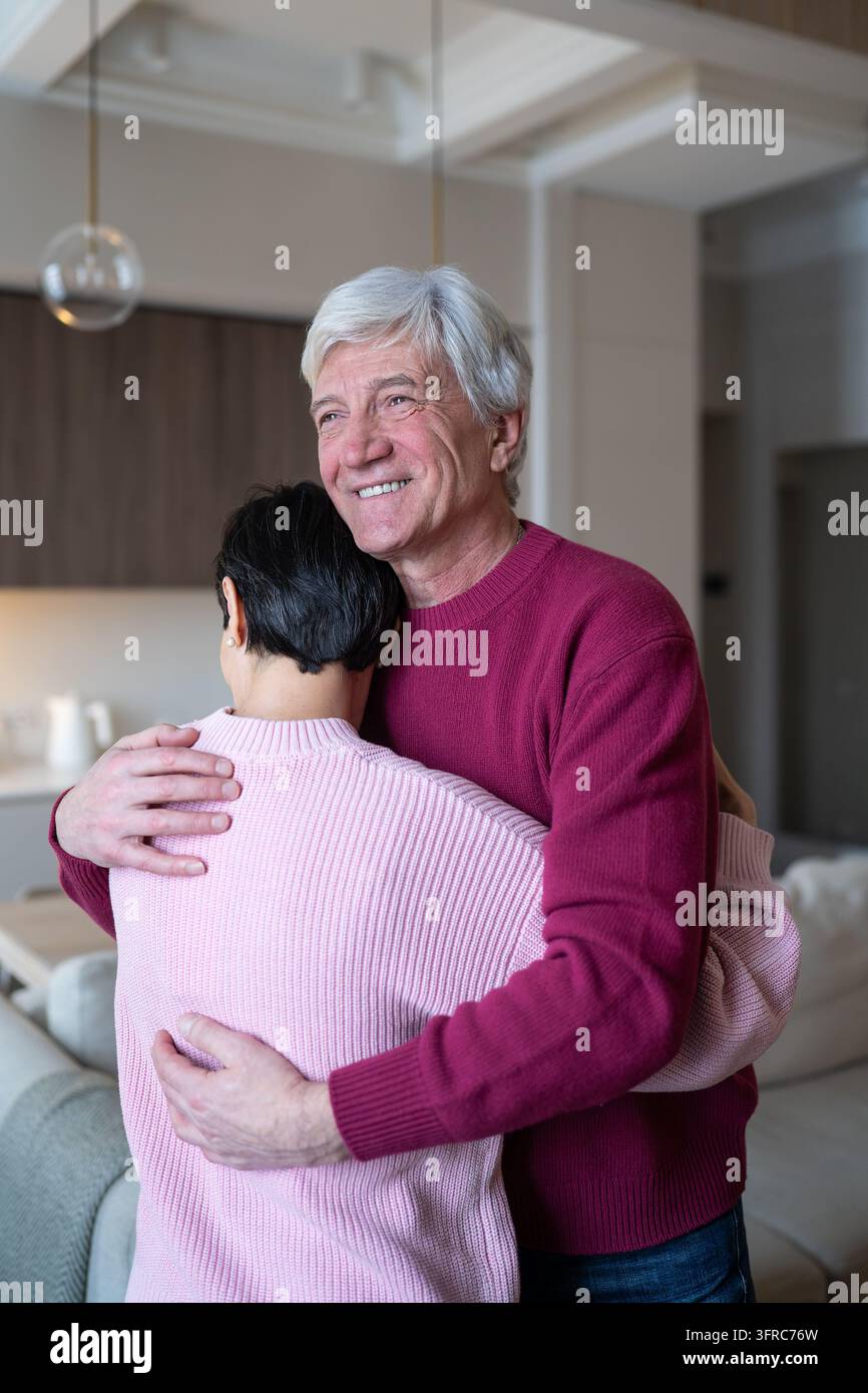 Marito anziano sorridente e felice che abbraccia con amore e affetto la sua amata moglie in soggiorno. Foto Stock