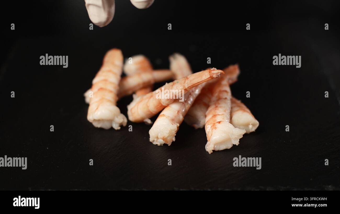 La mano guantata dello chef che fa cadere gamberetti cotti su un piatto di ardesia scura, preparando un piatto di frutti di mare Foto Stock