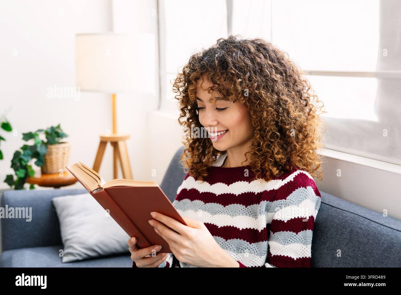 Donna sorridente che legge un libro avvolta in una coperta accogliente Foto Stock