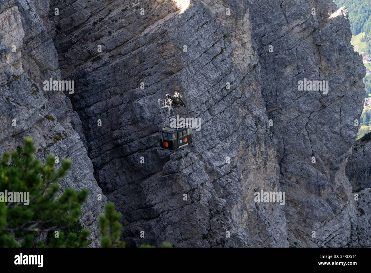 Una piccola funivia è sospesa a cavi d'acciaio mentre passa accanto a ripide e frastagliate pareti di roccia calcarea nelle Dolomiti. La luce del sole evidenzia la texture rocciosa Foto Stock