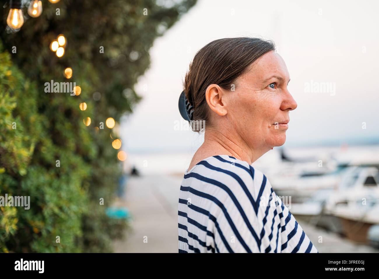 Donna pensierosa che guarda in lontananza vicino a un calmo porto marittimo all'alba. Concetto di riflessione, consapevolezza delle vacanze e momenti mattutini tranquilli. Sele Foto Stock