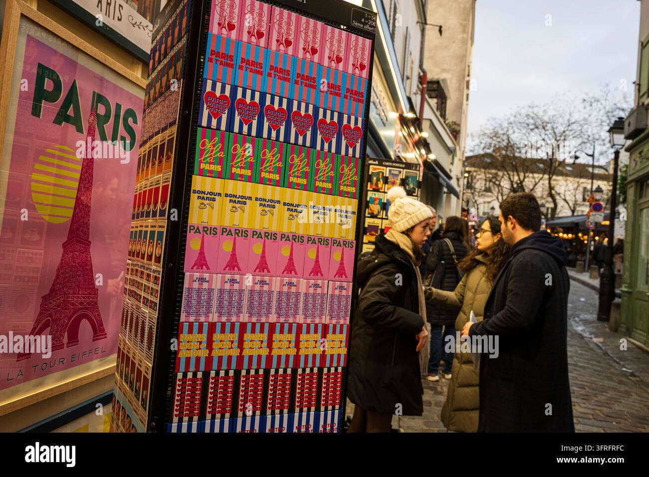 Parigi, Francia - 30 marzo 2026: Pedoni passano davanti a uno squisito espositore di souvenir pieno di magneti e poster colorati a tema Parigi lungo un Mo Foto Stock