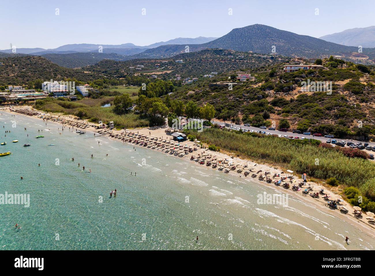 Vista aerea di Almyros Beach vicino ad Agios Nikolaos a Creta, che mostra una costa sabbiosa con ombrelloni, acqua di mare limpida e colline verdi circostanti Foto Stock