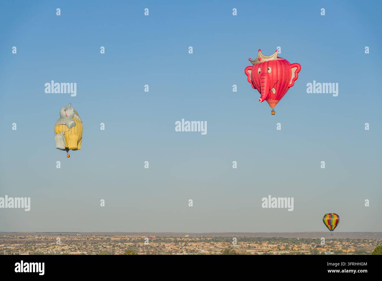 Due grandi mongolfiere a forma di elefante, una rosa con una corona e l'altra grigia e gialla, galleggiano in un cielo blu terso sopra una città estesa e un deserto Foto Stock