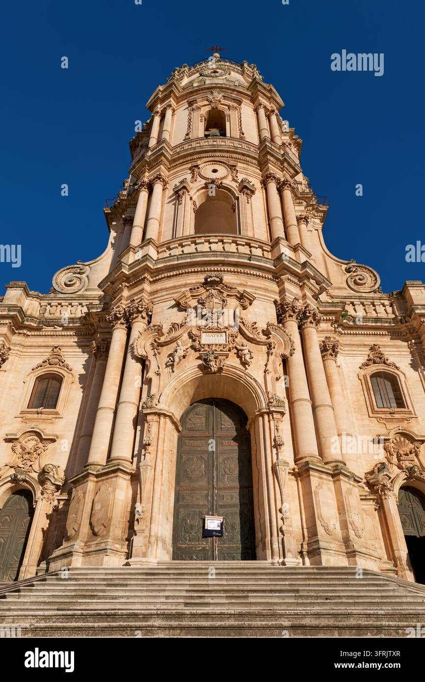 Facciata barocca della torre della Chiesa Madre di San Giorgio a Modica Alta, una città del Val di Noto inserita nella lista del Patrimonio Mondiale dell'UNESCO, vista dai gradini sotto un cielo blu terso Foto Stock