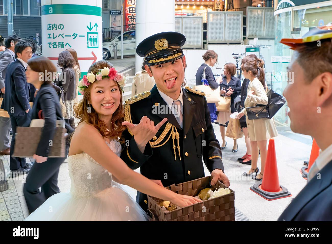 Sposini in abito bianco e uniforme cerimoniale che distribuiscono regali da un cesto agli invitati di nozze su una strada serale affollata a Tokyo Foto Stock