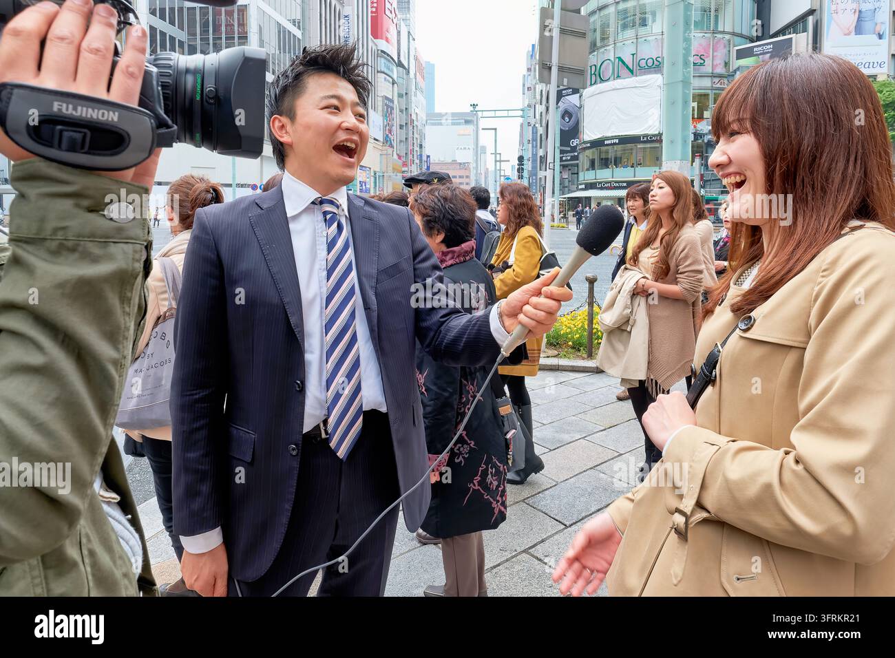 Un reporter che tiene un microfono intervista una giovane donna che ride mentre un operatore di telecamera filma su un marciapiede affollato nel distretto commerciale di Ginza Foto Stock