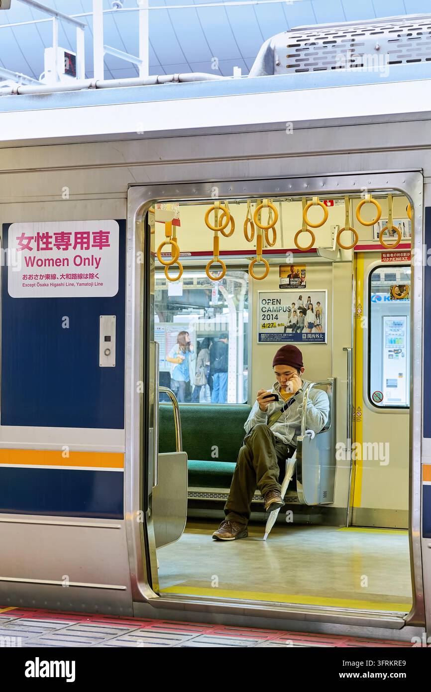 Un passeggero maschile guarda un telefono cellulare sulla panca di un vagone del treno pendolare contrassegnato "Solo per donne", visto dal marciapiede in Giappone Foto Stock