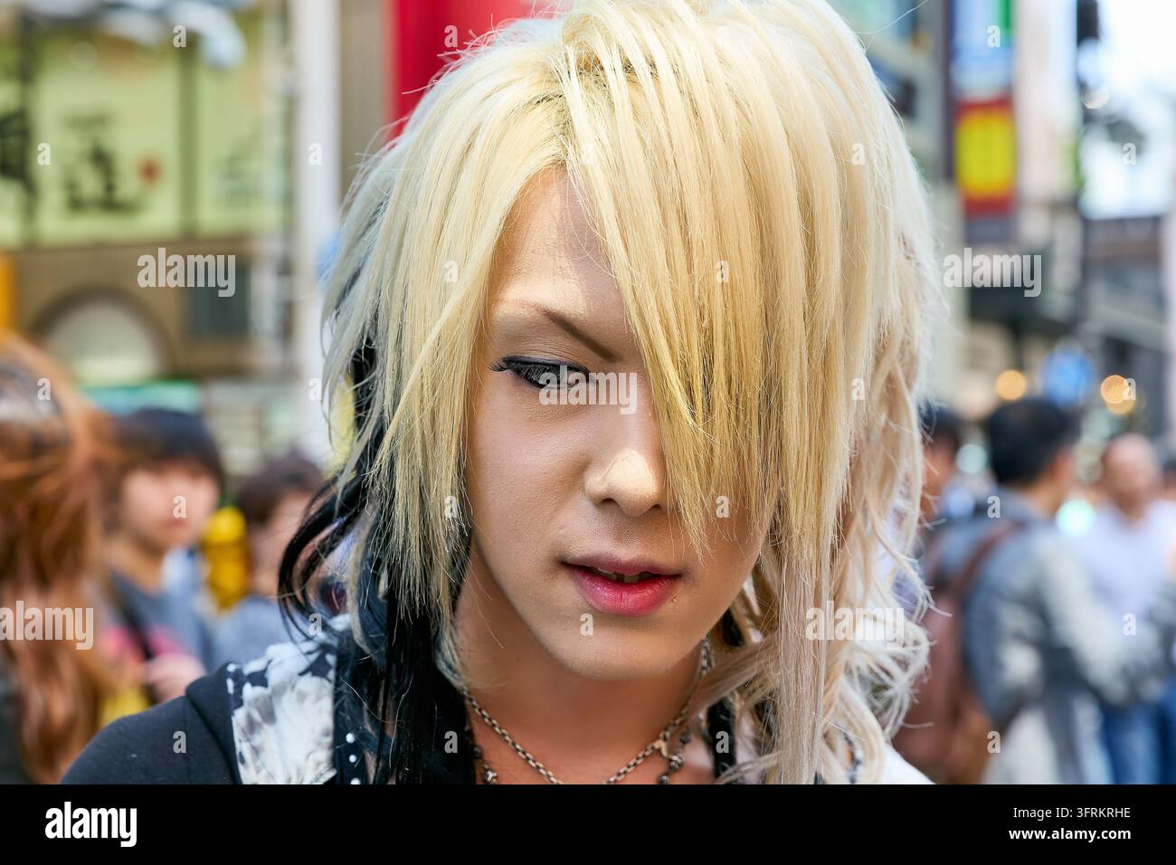 Prima di un giovane adulto con trucco in stile visual kei, eyeliner e ciocche tinte di nero, tra una folla animata diurna a Osaka, Giappone Foto Stock