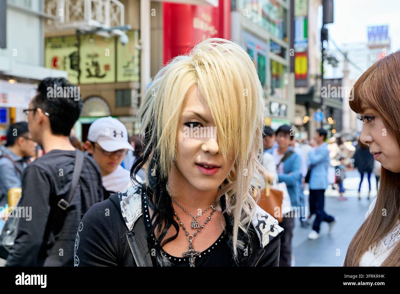 Giovane con capelli biondi decolorati, trucco occhi e collana con pendente a croce si trova tra i pedoni in un vivace quartiere commerciale di Osaka, Giappone. Foto Stock