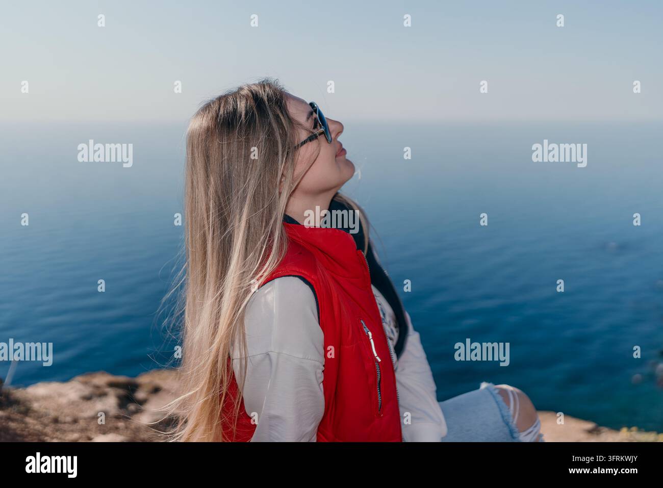 Donna seduta su una scogliera oceanica indossa un giubbotto rosso in un punto panoramico costiero guardando il cielo Foto Stock