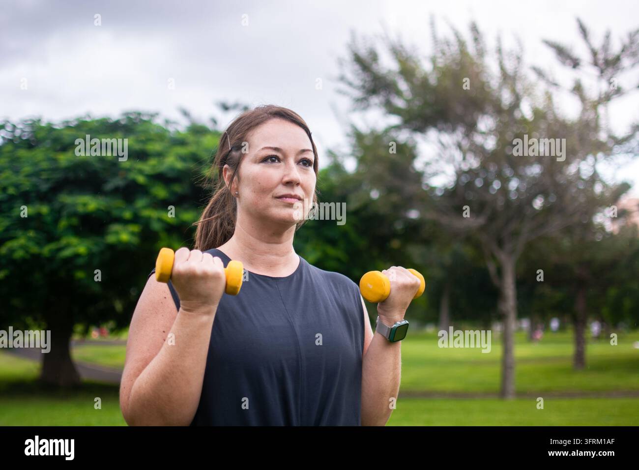 Donna in abbigliamento sportivo che esegue curl bicipiti con manubri gialli durante l'allenamento all'aperto in un parco pubblico verde, concentrandosi sulla forza e sulla fisi Foto Stock