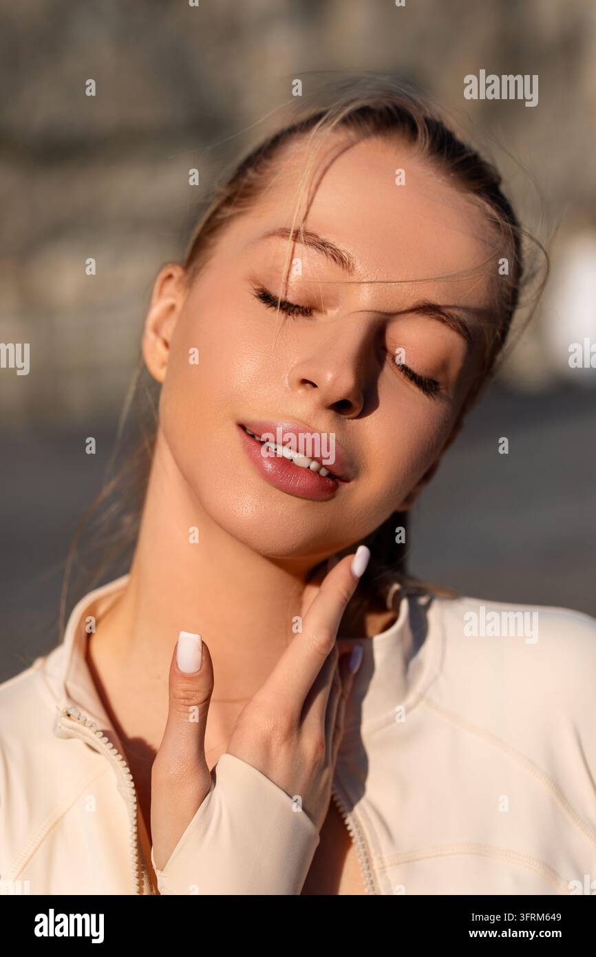 Donna attraente che accarezza dolcemente una carnagione luminosa con grazia. Donna di bellezza che presenta una bellezza facciale fresca sotto la luce del sole. Concetto di bellezza. Foto Stock