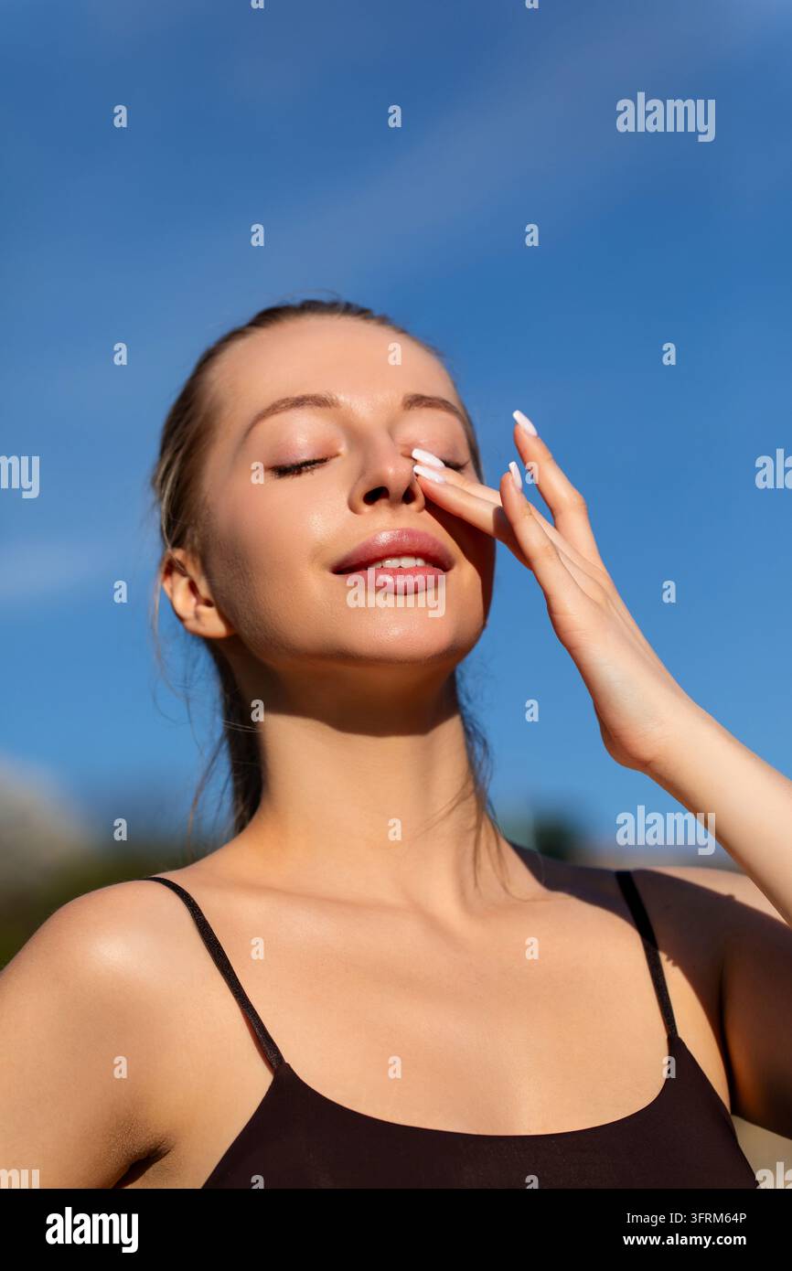 Donna attraente che accarezza dolcemente un incarnato luminoso con grazia. Donna di bellezza che presenta una bellezza facciale fresca sotto il cielo blu. Concetto di bellezza. Foto Stock