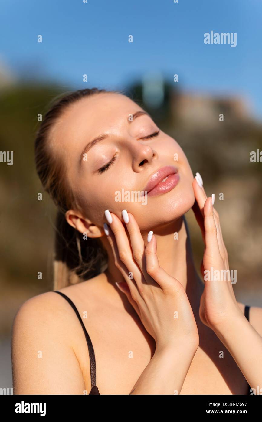 Bella donna accarezza dolcemente un viso luminoso pacificamente. Donna attraente mostra una pelle fresca e liscia naturalmente all'aperto. Concetto di bellezza. Foto Stock