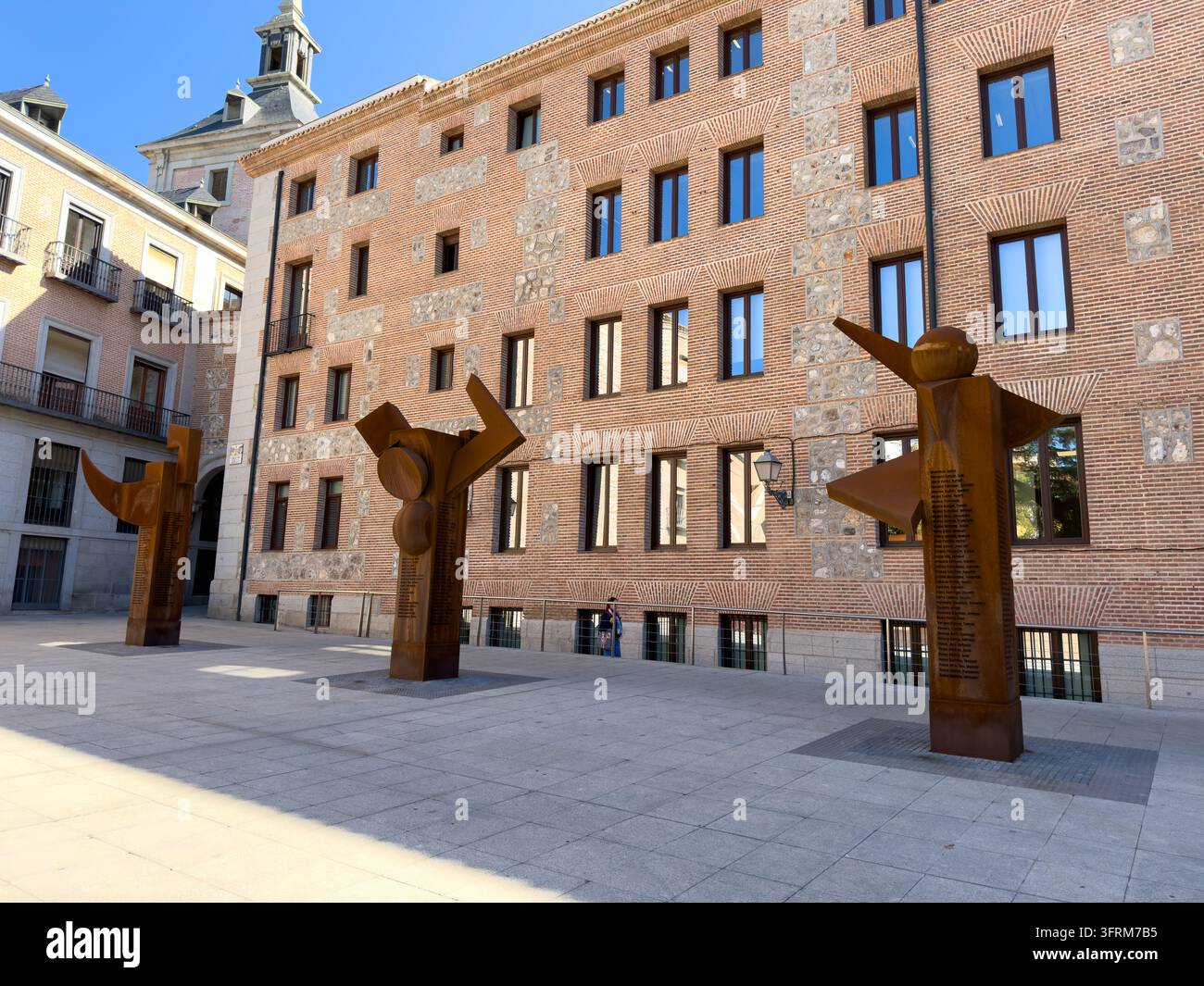 MADRID, SPAGNA - 09 OTTOBRE 2026 Figure metalliche scultoree occupano un cortile lastricato di Madrid accanto all'architettura storica in mattoni. Forme angolari color ruggine Foto Stock