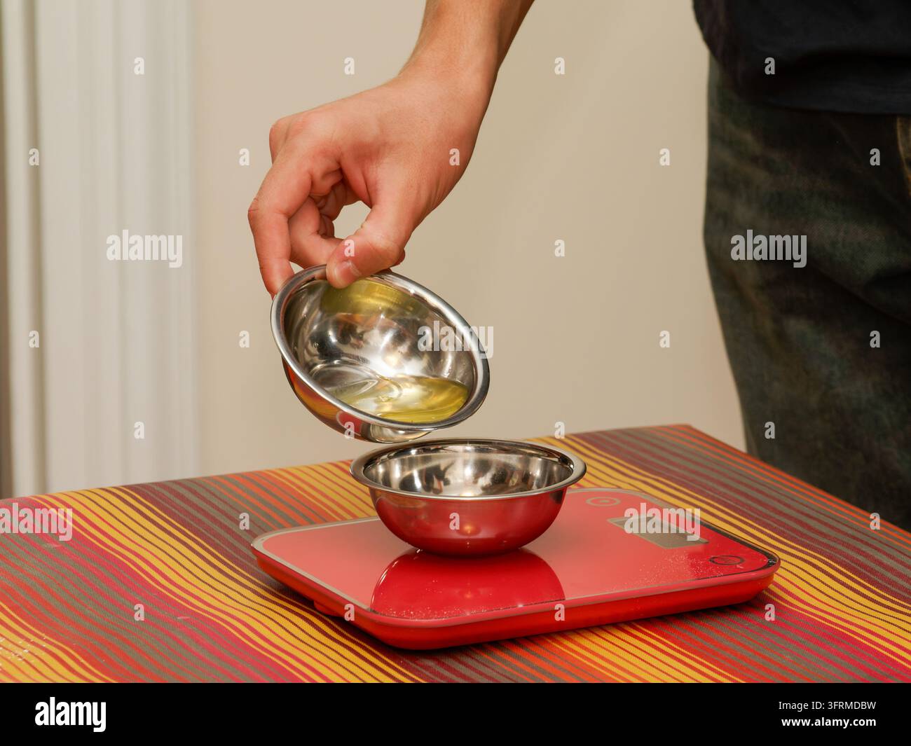 Persona che versa un liquido da una ciotola di metallo a un'altra su una bilancia da cucina digitale rossa. Foto Stock