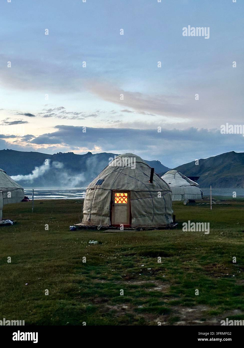 Focolare e casa: una porta illuminata e il fumo del camino che si arriccia danno vita a un campo di yurte sul fiume vicino al lago Kel-Suu, in Kirghizistan Foto Stock