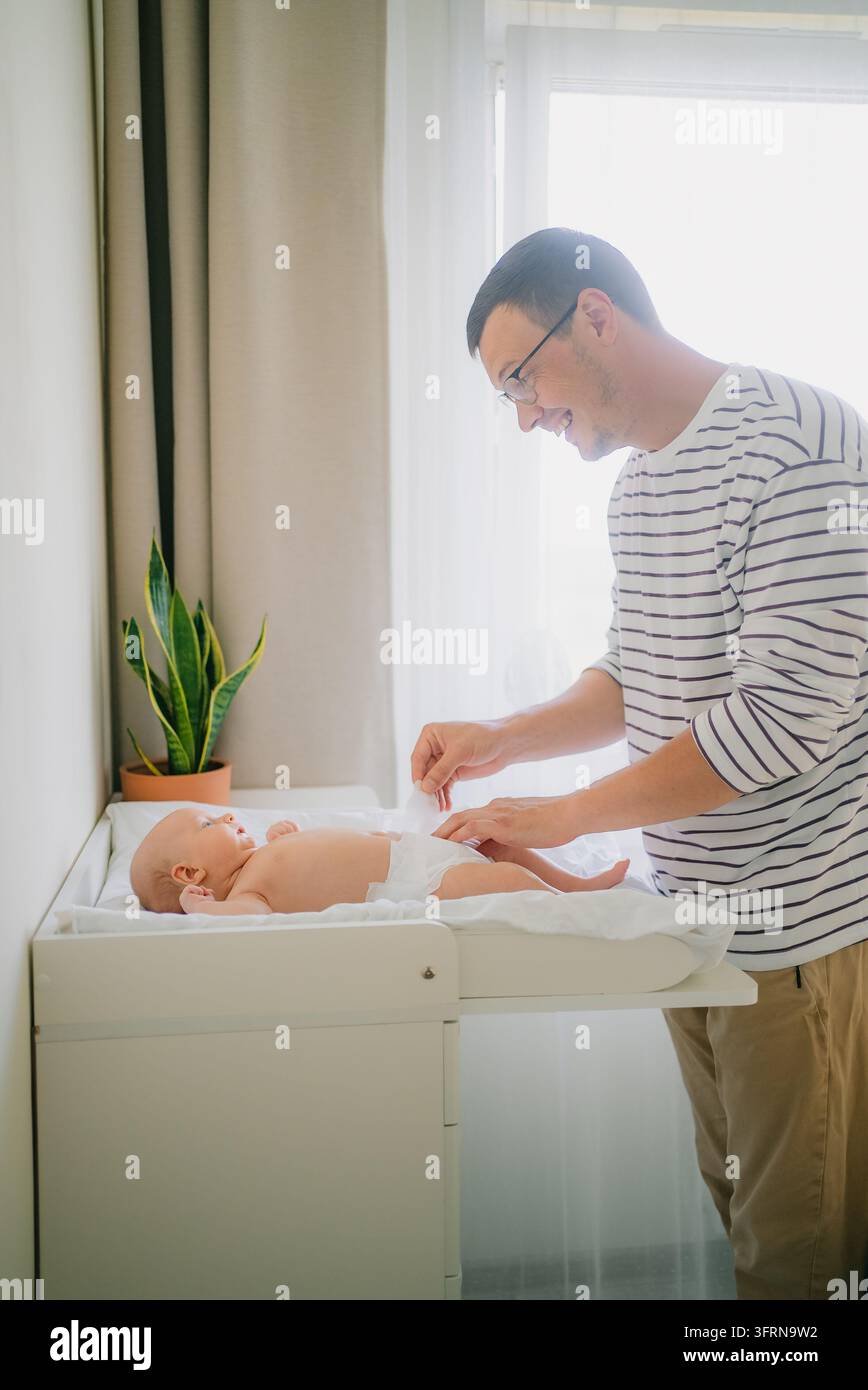 Padre che si prende cura del suo bambino, cambia il pannolino su un tavolo da cambio in una stanza luminosa, mostrando un momento d'amore del legame genitoriale e della routine familiare quotidiana Foto Stock