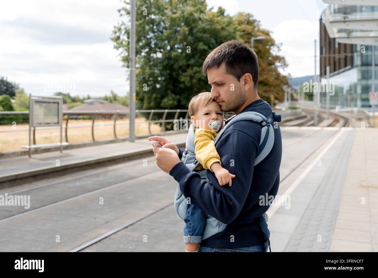 Padre che porta il suo bambino in un marsupio davanti a sé mentre aspetta accanto ai binari del tram alla fermata della Luas di Central Park a Dublino, Irlanda, per il trasporto pubblico urbano Foto Stock