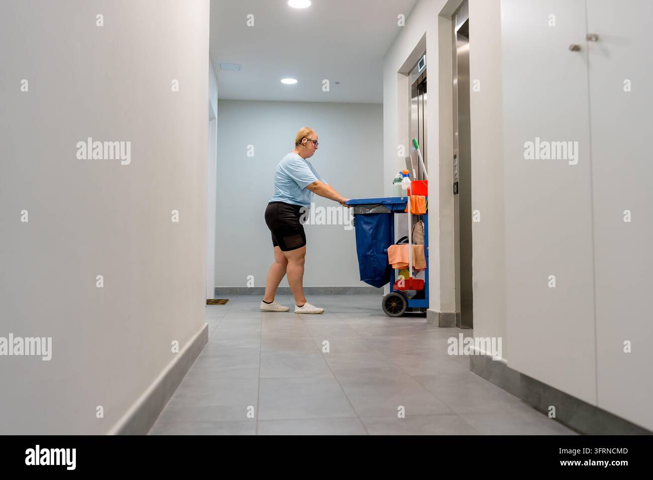 Addetto alle pulizie spinge un carrello per le pulizie attraverso un corridoio di un appartamento moderno, portando materiale e attrezzature per la sanificazione e la manutenzione di routine Foto Stock