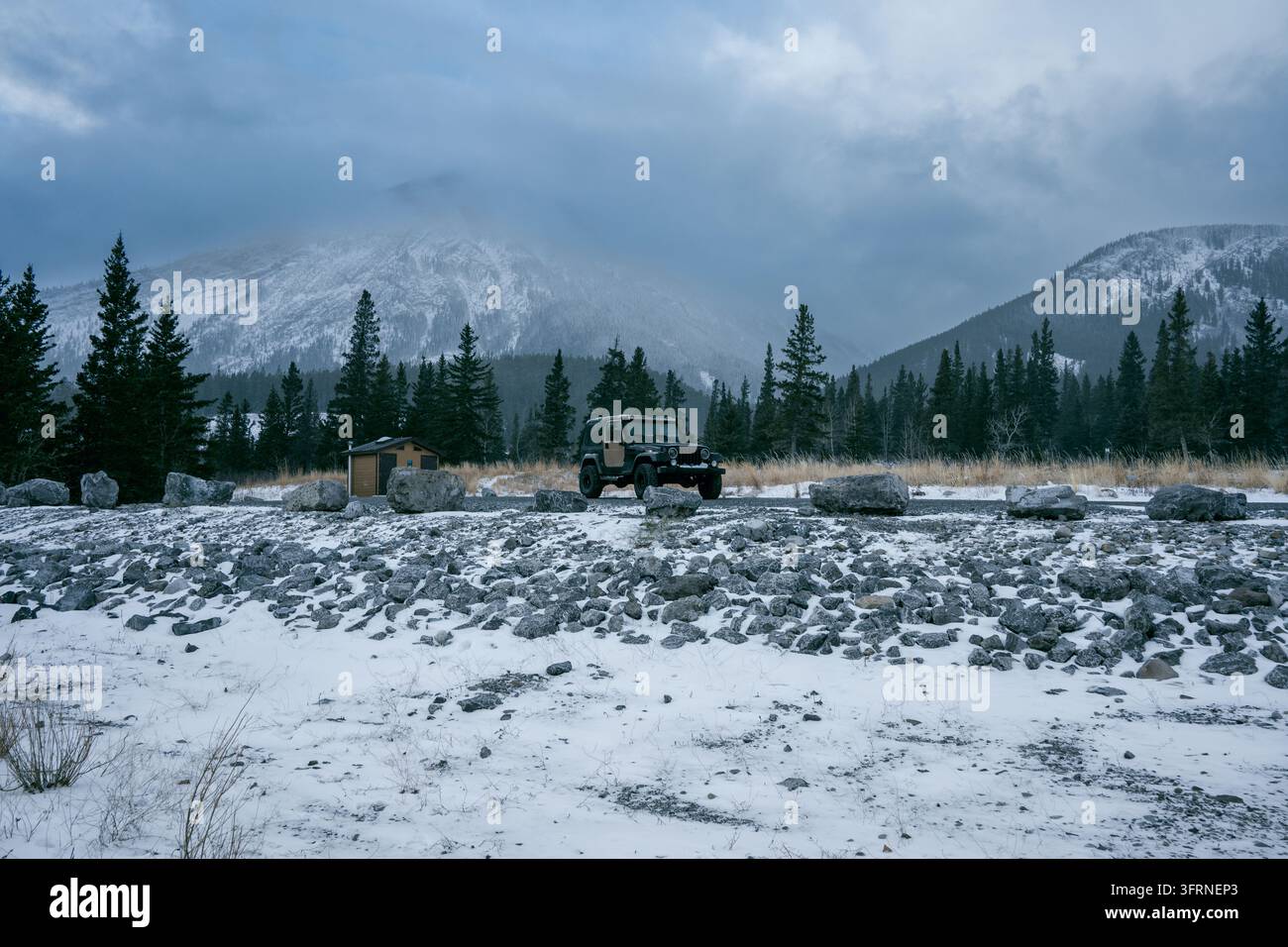 Inquadratura ampia all'altezza degli occhi di un veicolo fuoristrada 4x4 nero parcheggiato accanto a una strada sterrata innevata vicino al lago Abraham, in Alberta, con foresta di pini, capanna e cielo nuvoloso Foto Stock