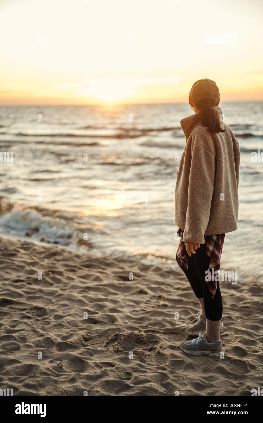 Donna in abiti sportivi casual osserva il tramonto dorato sul mare mentre è in piedi su una spiaggia sabbiosa, con onde gentili e un orizzonte calmo della sera. Foto Stock